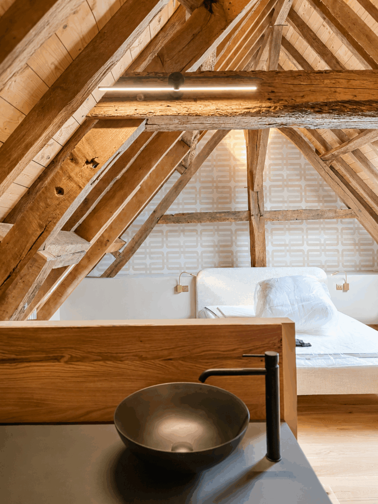 Chambre aménagée sous une charpente ancienne rénovée, illustrant la transformation d'un bâtiment existant et la valorisation du patrimoine par TFL GROUPE.